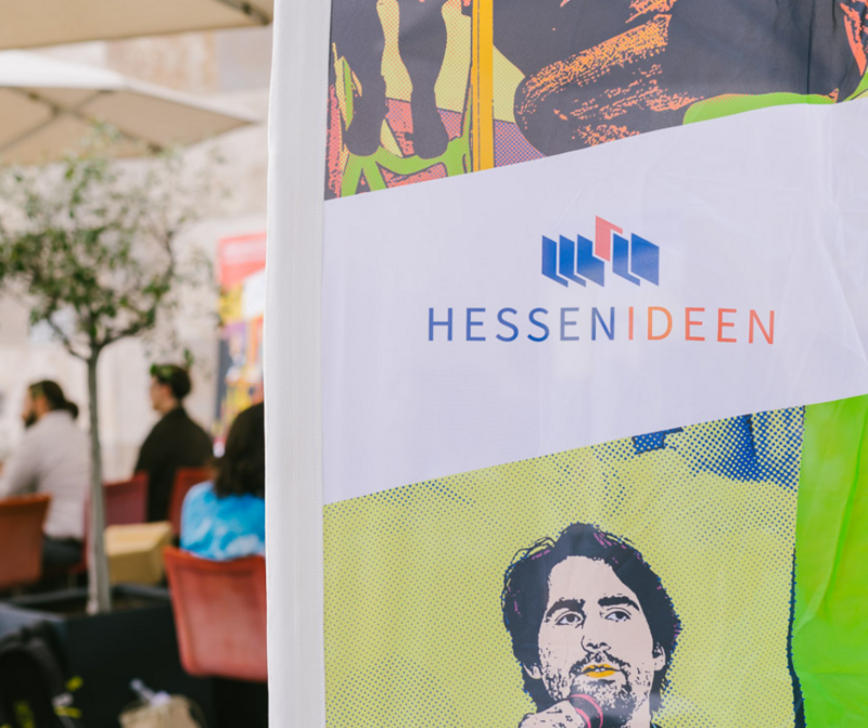 Links auf dem Bild erkennt man einen unscharfen Hintergrund. Der Hintergrund zeigt einen Raum, in dem ein kleiner Baum steht und Menschen auf roten Stühlen sitzen. Rechts auf dem Bild erkennt man einen Header, auf dem Hessen Ideen steht. Der Header ist bunt und bildet den Kopf eines Mannes, der am Mikrofon steht ab. 