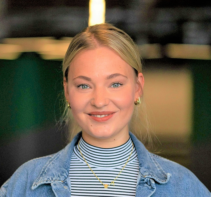 Auf dem Foto erkennt man eine junge blonde Frau. Sie trägt eine Jeansjacke und einen schwarz-weiß gestreiften Rollkragenpullover darunter. Zudem trägt sie goldene Ohrringe und eine goldene Kette. Sie hat blaue Augen und lächelt freundlich in die Kamera.